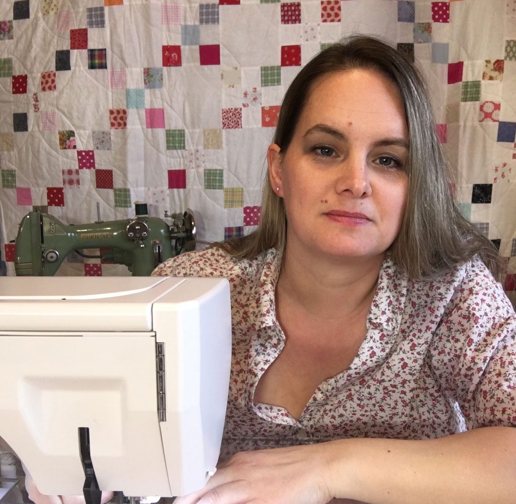 Caroline at her sewing machine, with a colorful quilt of small squares hanging behind her.