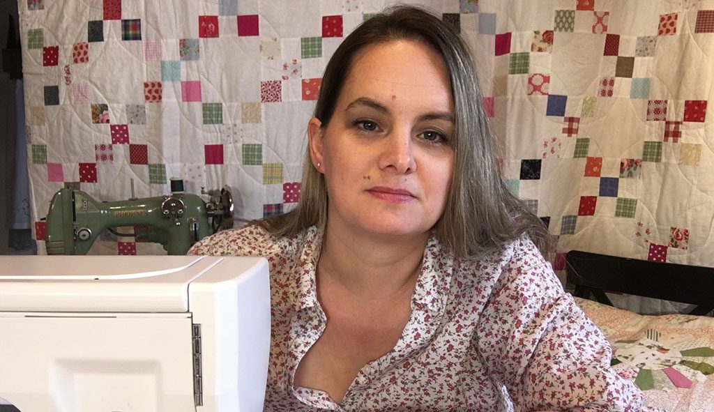 Caroline sitting at her sewing machine and looking into the camera, with a quilt of small colorful squares behind her.