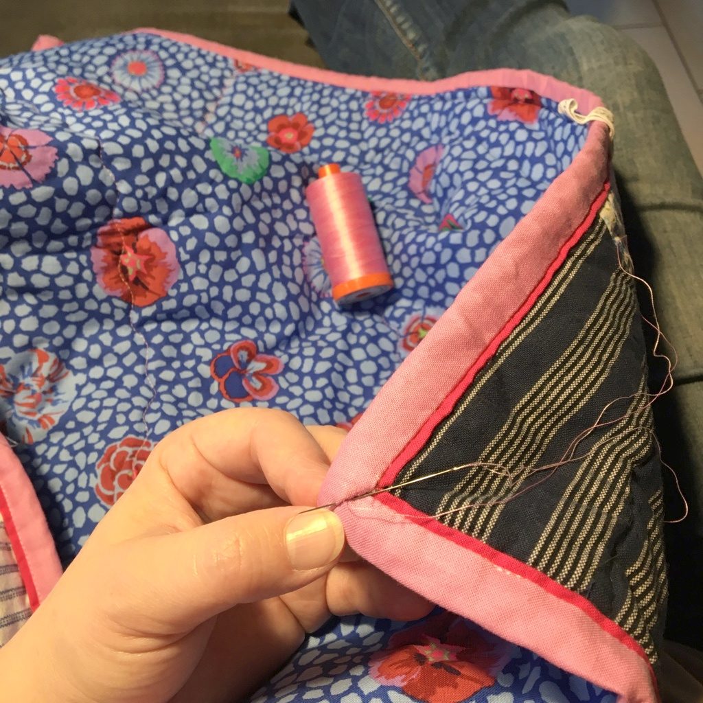 Carolines Hand näht das rosafarbene Binding an Evas Quilt von Hand fest.