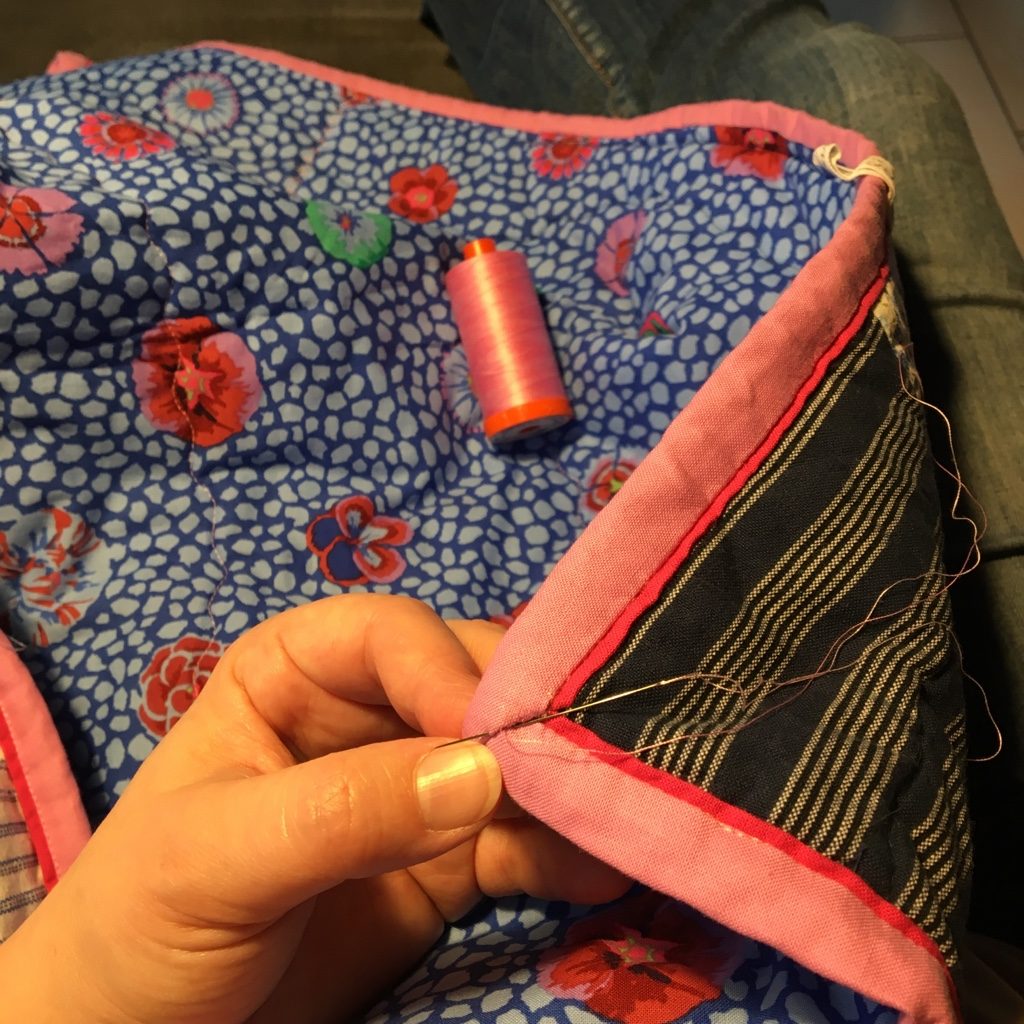 Caroline’s hand stitching a pink binding with a narrow red edge onto a blue floral quilt, a spool of pink thread beside it.