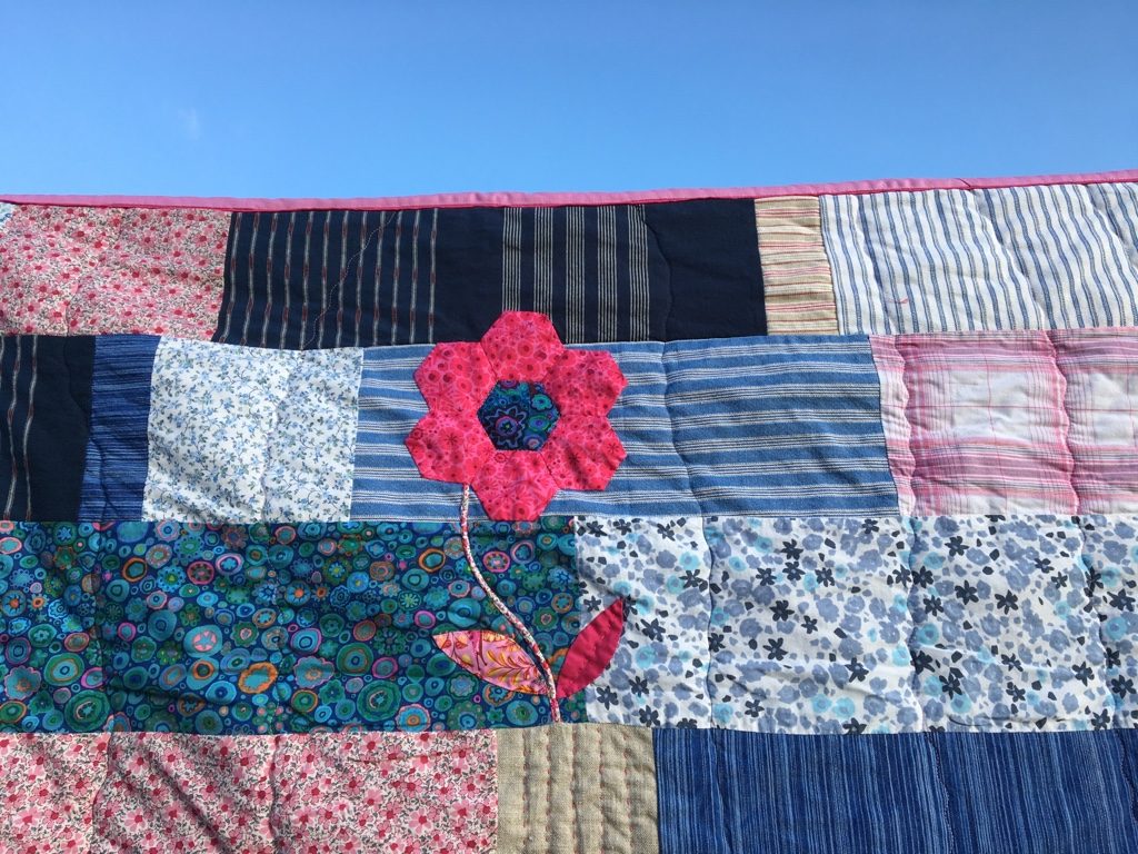 Detail of Eva’s Quilt: a pink flower with a stem of Liberty fabric, appliquéd onto patchwork of blue shirt and blouse fabrics.