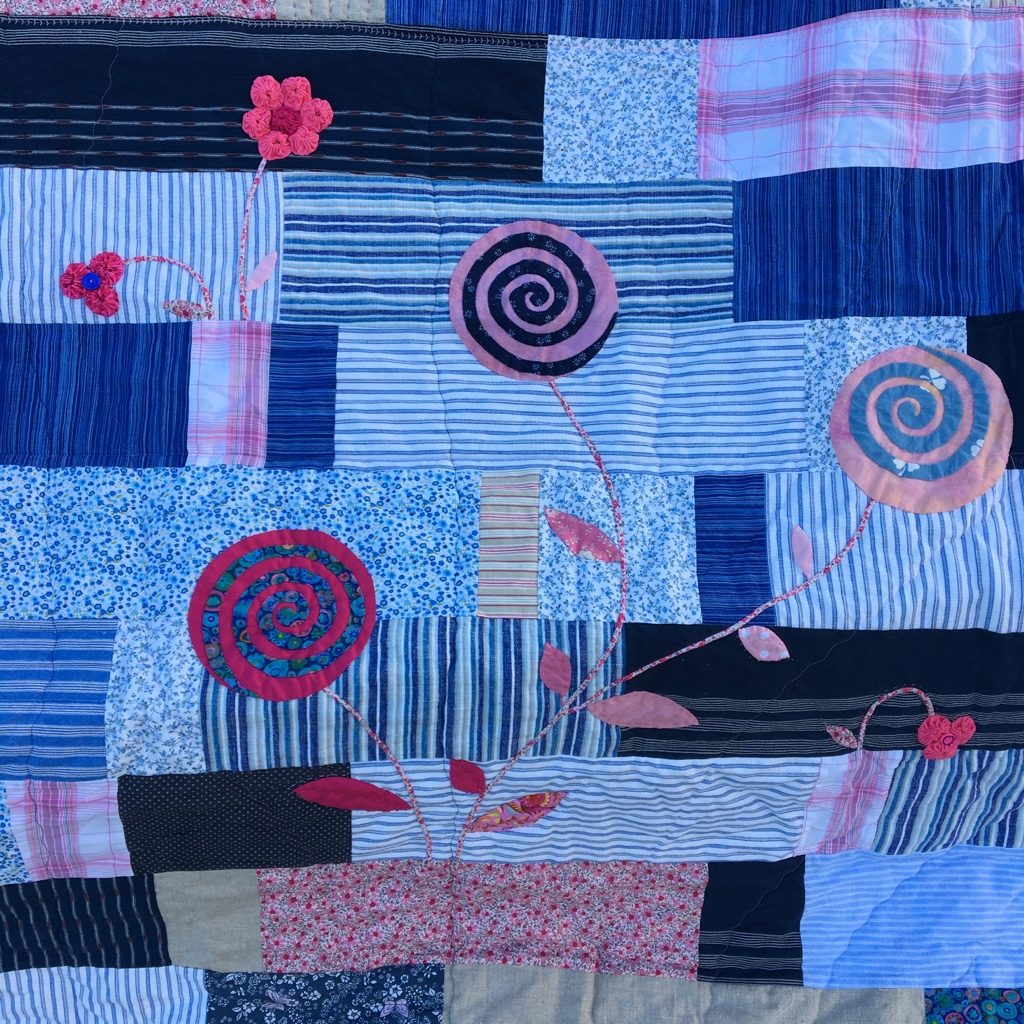 Eva’s Quilt laid out flat: strips of blue shirt and blouse fabrics, with spiral flowers in blue and pink and long-stemmed flowers in between.