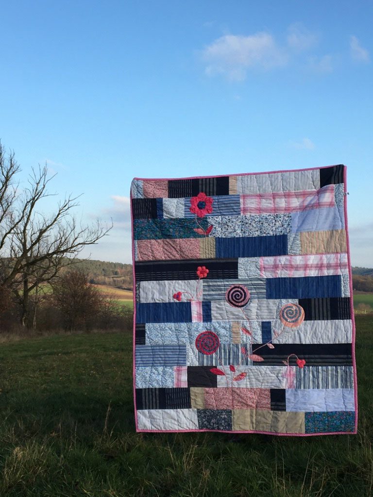 Eva’s Quilt hanging in a meadow against fields in the evening light, a quilt of blue and pink fabrics with appliquéd flowers.