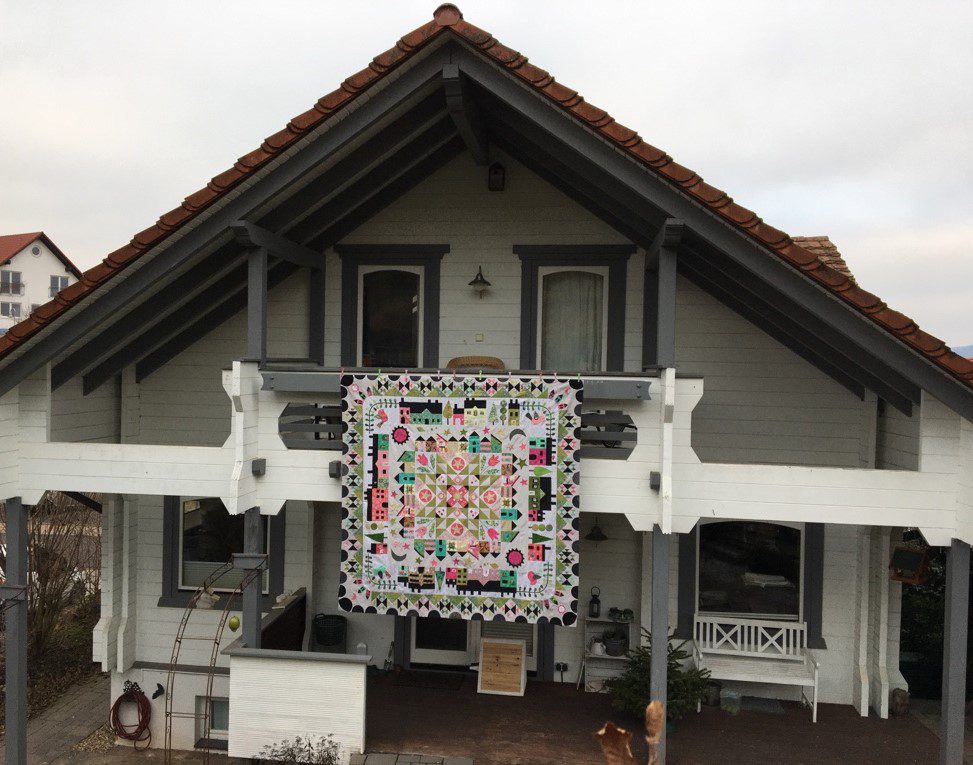 The white-painted timber house with a dark roof, a colorful quilt hanging from the balcony and a garden in front.