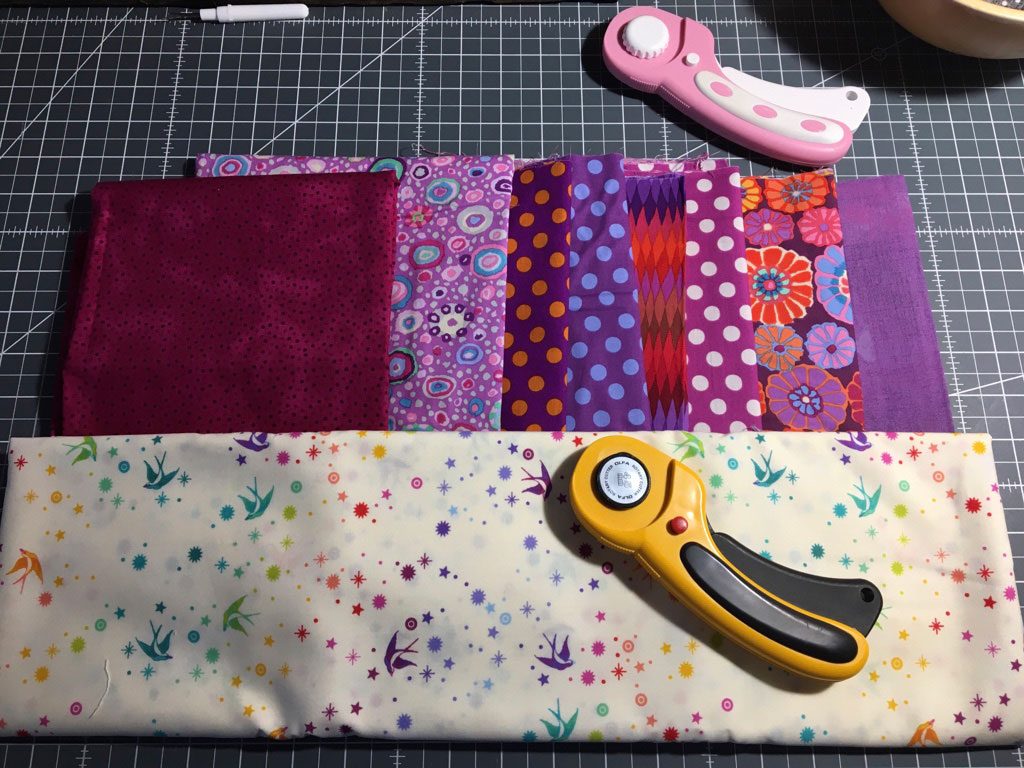 Purple and pink fabrics on a cutting mat, with a light fabric with colorful dots and a rotary cutter in front.