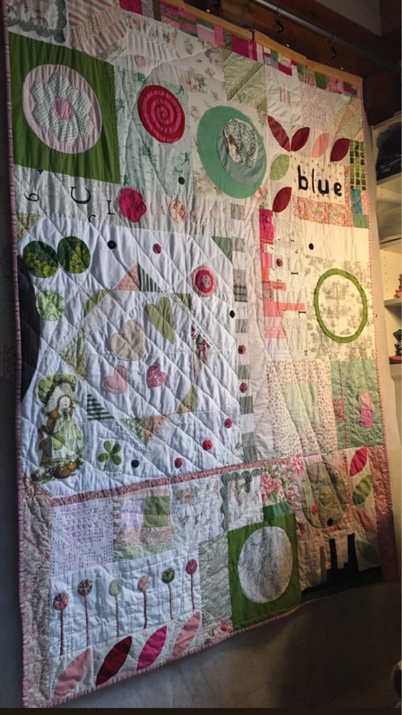 The Vignettes Quilt hanging on a wall in half-light, with light falling across the quilting stitches.
