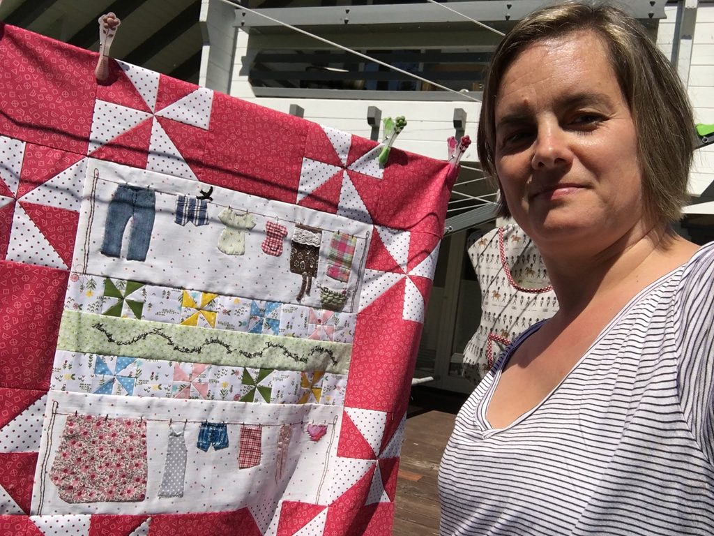 Caroline holding up her wall quilt Windy Washday, with laundry on a clothesline, for the camera. She is wearing a striped shirt.