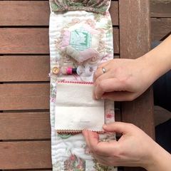Two hands open a handmade sewing roll with a pincushion and pockets on a wooden table.