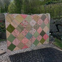 A quilt of on-point squares in pink and green hangs on a line in the garden above paving stones.