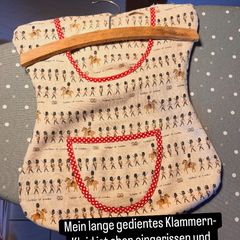 A dress-shaped clothespin bag in fabric with little soldiers and red polka-dot trim, on a wooden hanger.
