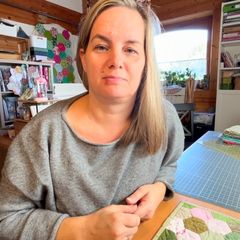 Caroline sits at her sewing table with fabric hexagons and looks into the camera.