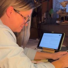 Caroline, wearing glasses, works on a tablet in her sewing room.