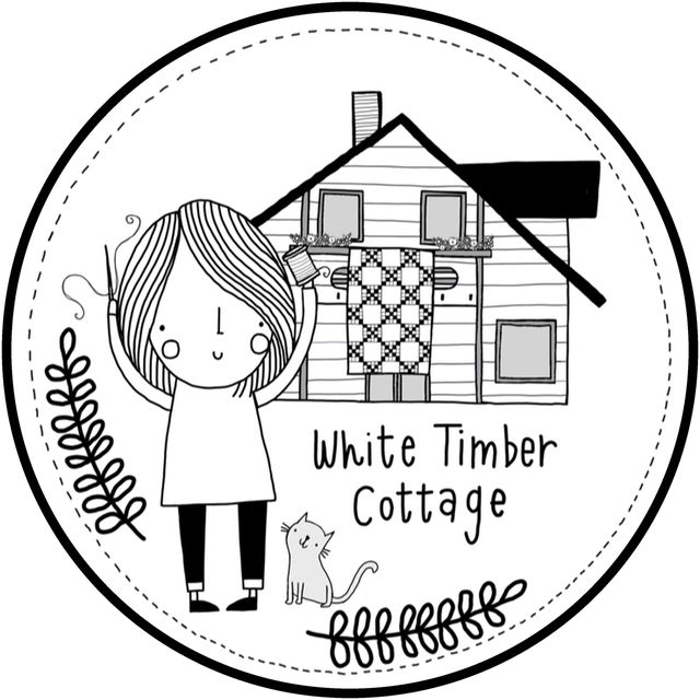 The White Timber Cottage logo as a round white sticker with a black border: Caroline with a needle and a spool of thread in front of her white timber house with a quilt at the window, a cat, sprigs and a circle of running stitches.